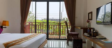 A hotel room with a bed, a desk, a chair, a TV, and a balcony with a view of greenery.