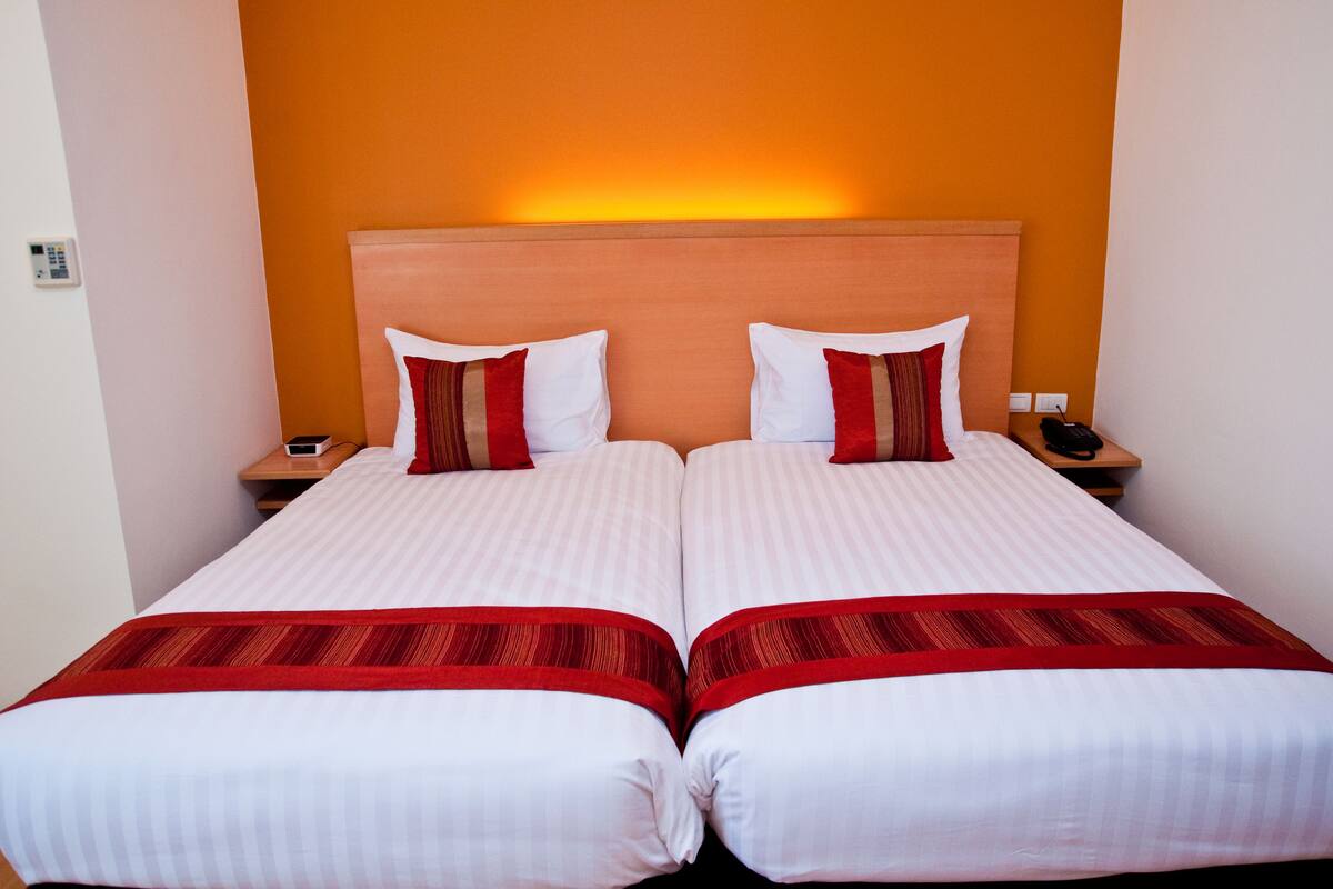 A double bed with two pillows and a red accent pillow, a bedside table with a phone, and a wall-mounted light.