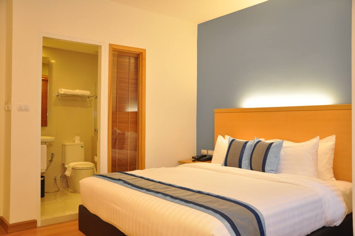 A hotel room with a bed, a nightstand, a bathroom with a toilet and sink, and a wooden door.
