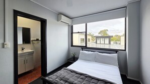 A bedroom with a bed, a window with a view of buildings, a bathroom with a sink and mirror, and an air conditioning unit.