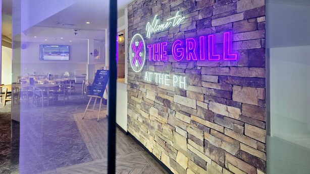 A restaurant interior with a stone wall and neon sign reading "Welcome to THE GRILL AT THE PH".
