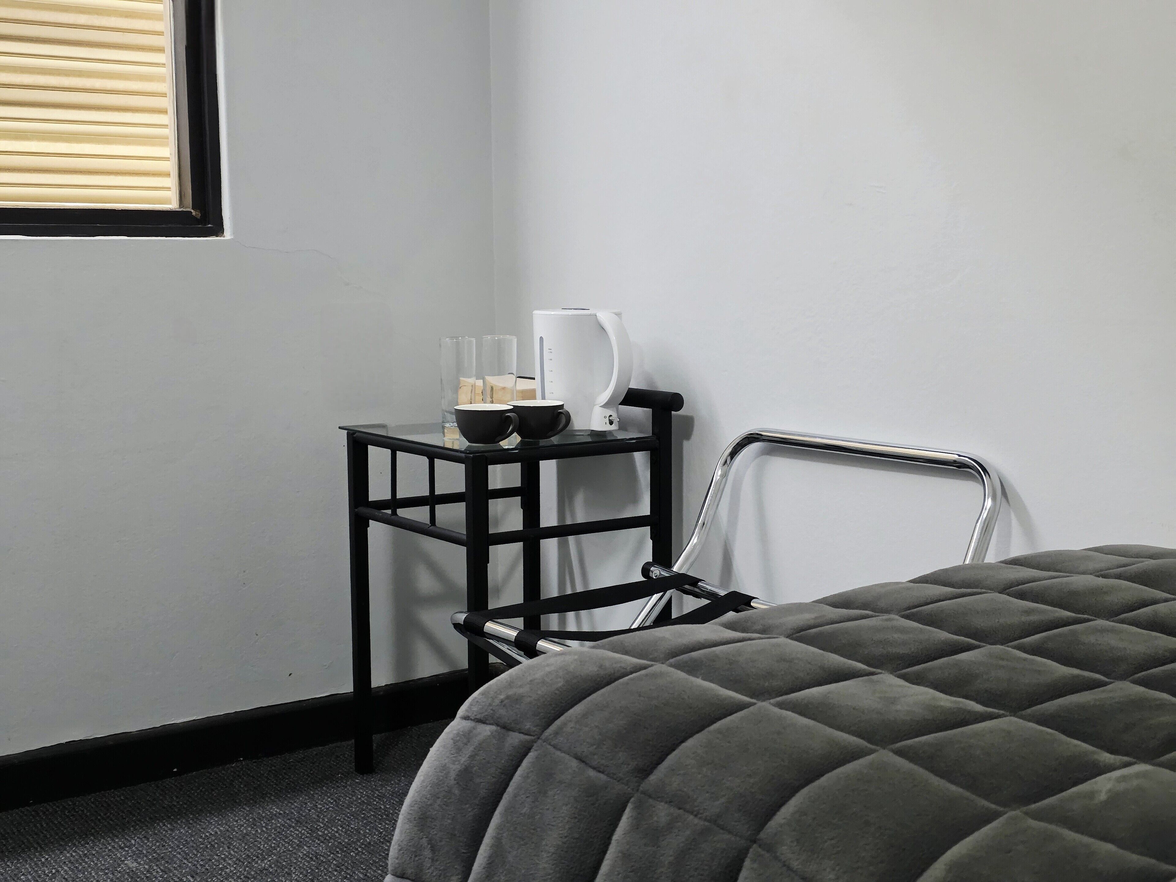A room with a bed, a small black table with a white kettle, two glasses, and a bottle.