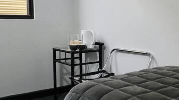 A room with a bed, a small black table with a white kettle, two glasses, and a bottle.