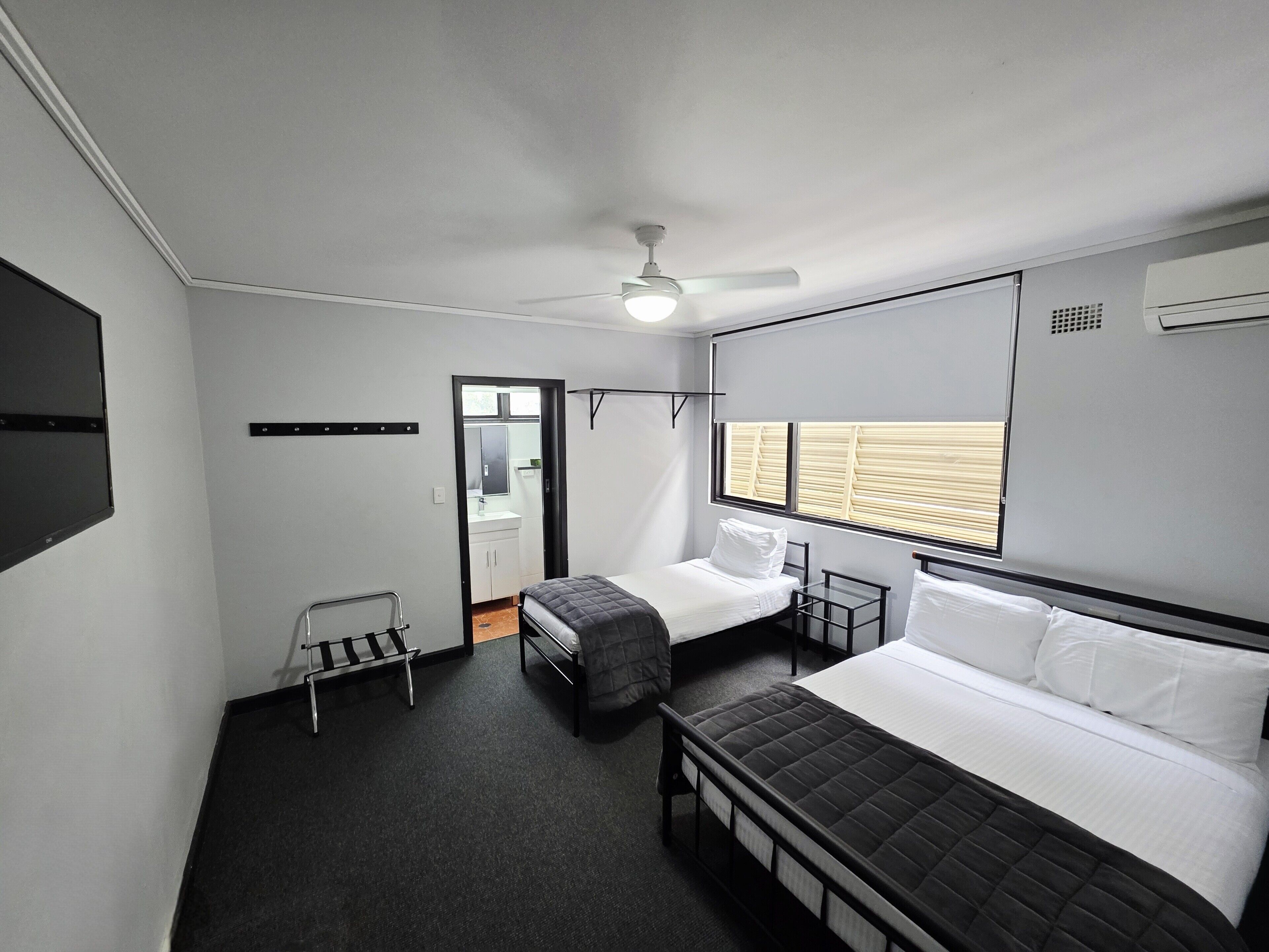 Ein kleines Schlafzimmer mit Bett, Nachttisch, Stuhl, einem Fenster mit Jalousien und einem Deckenventilator.