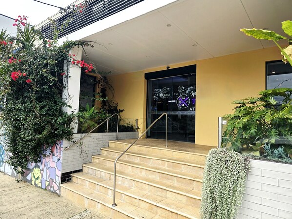 A building entrance with a staircase leading to a glass door, surrounded by greenery and a mural. - Panania Hotel (Panania)