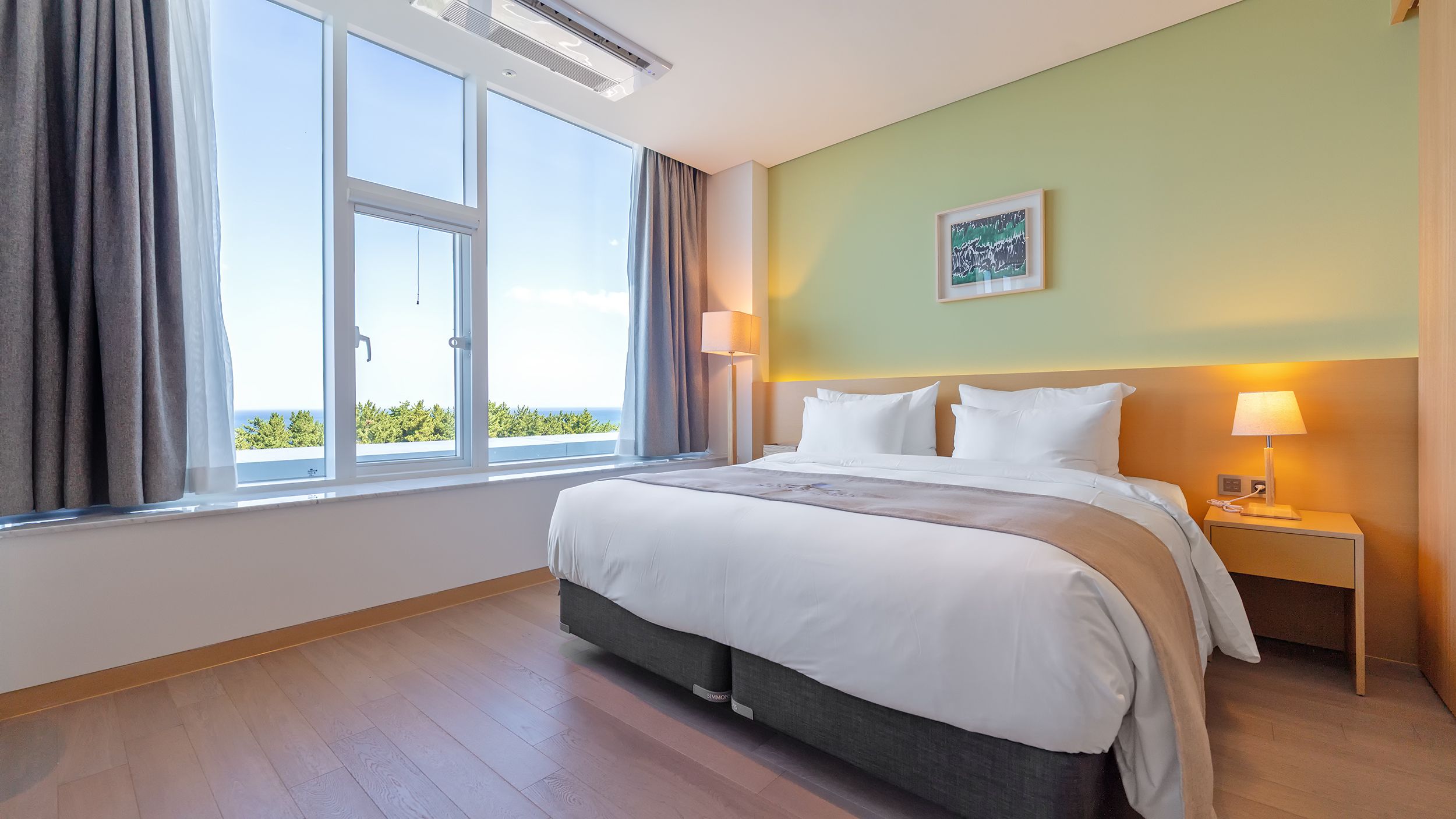A hotel room with a large window, a bed with white linens, a nightstand, and a lamp.