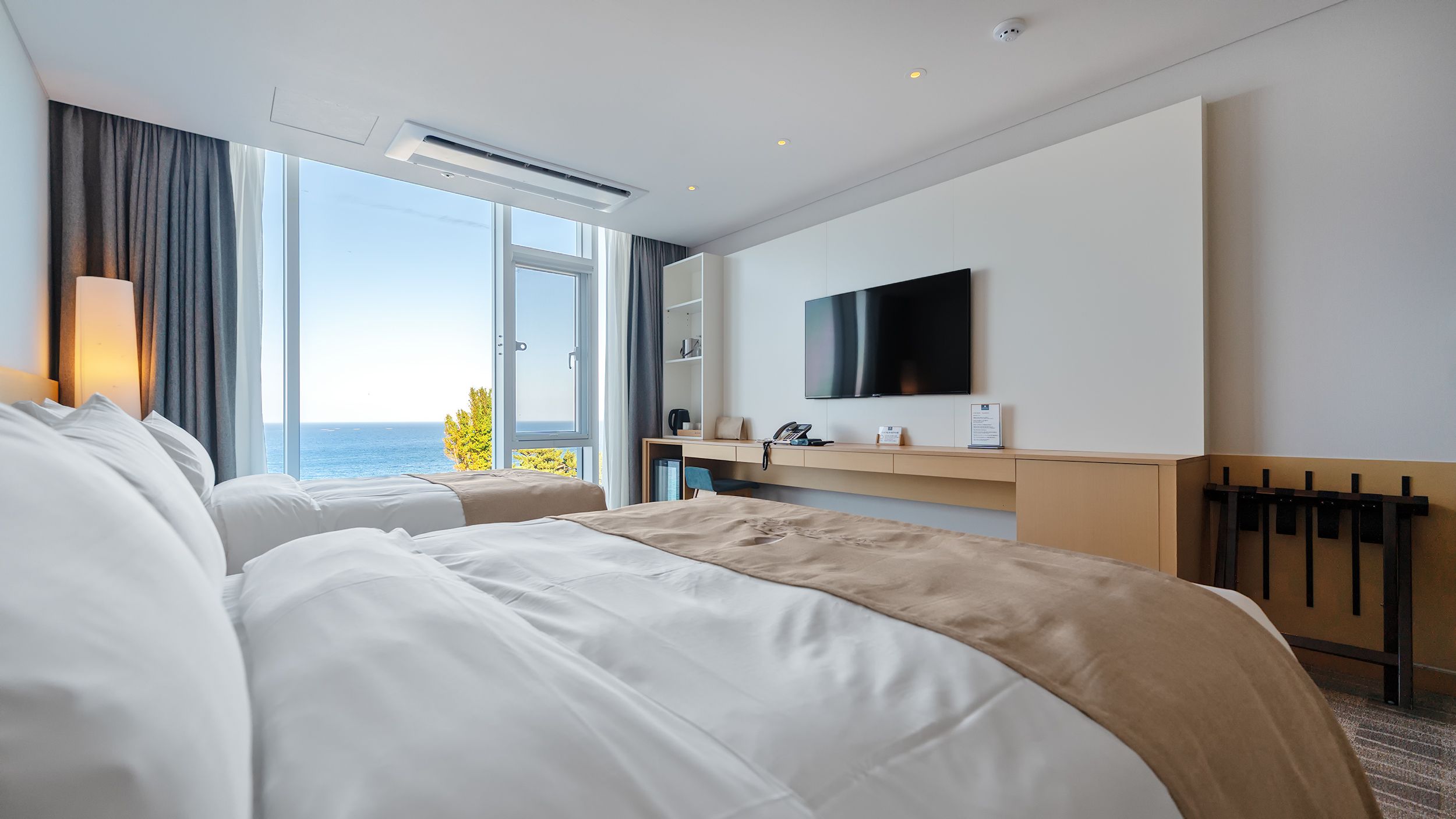 A hotel room with two beds, a TV, a desk with a phone, and a view of the sea.