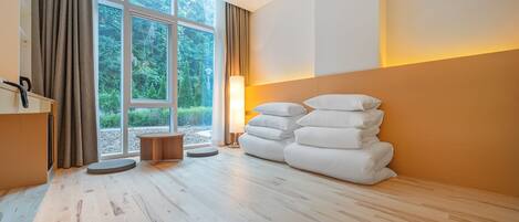 A room with a large window, a bed with white pillows, a standing lamp, and a small wooden table.