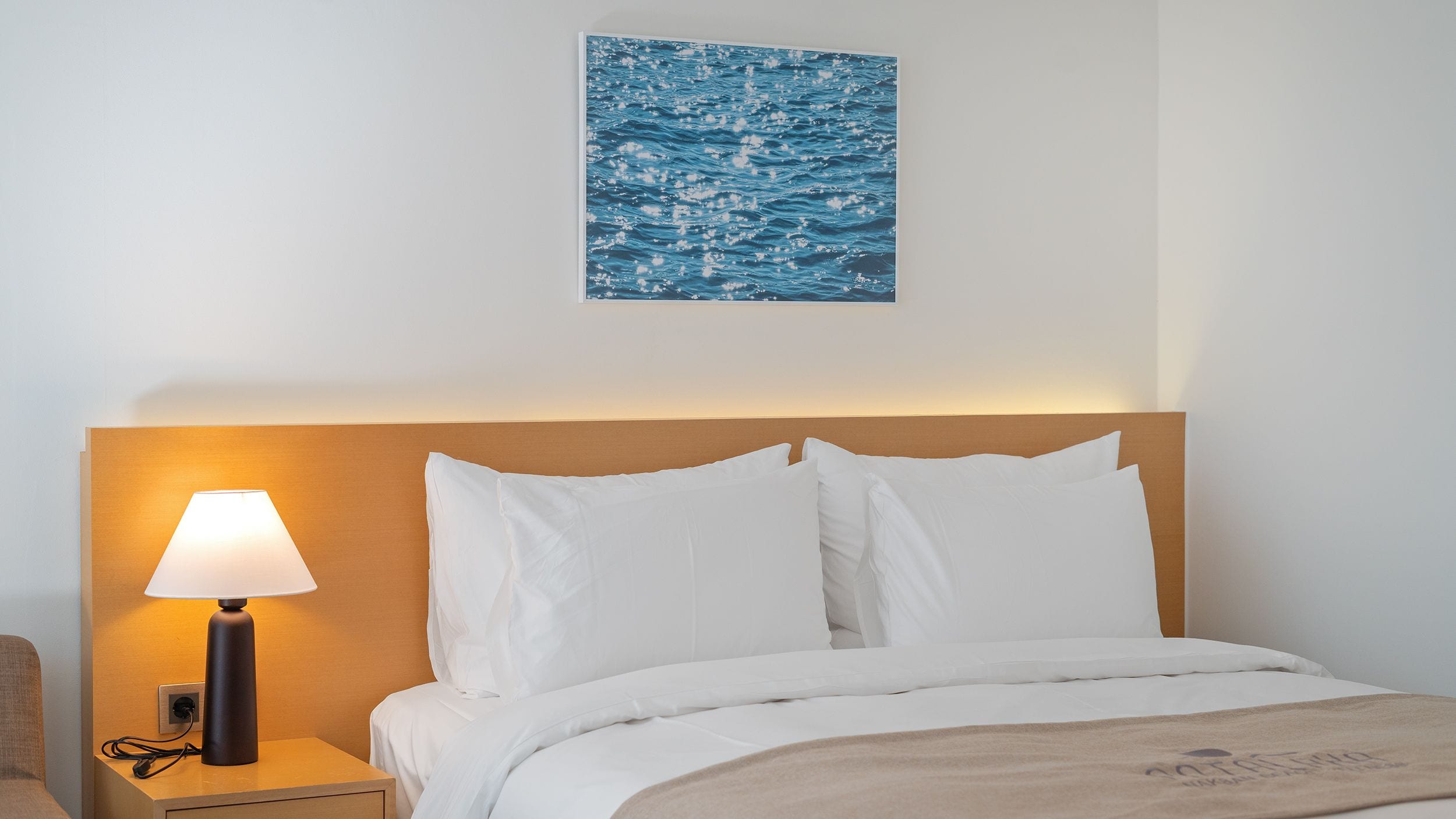 A hotel room with a bed, white bedding, a nightstand with a lamp, and a wall-mounted artwork.