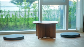 Une petite table en bois, accompagnée de deux tabourets ronds, est placée devant une fenêtre donnant sur un paysage verdoyant.