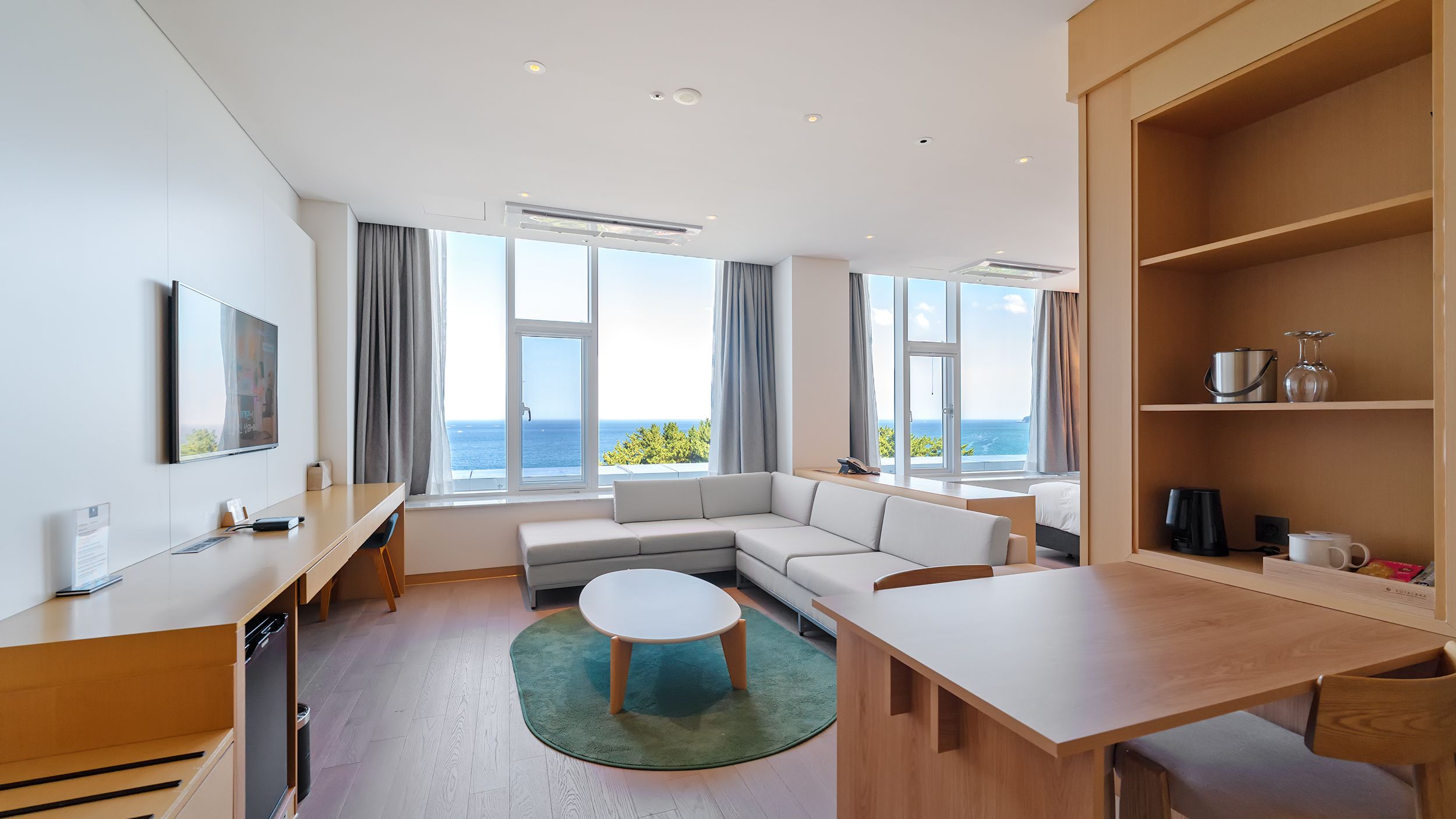 A modern hotel room with a large window offering a view of the sea, a sofa, a dining table, and a small kitchen area.