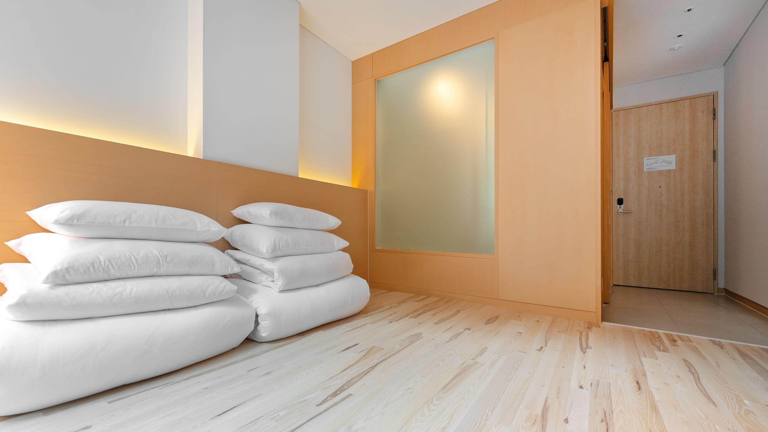 Une chambre d’hôtel avec un plancher en bois, une pile d’oreillers blancs, une porte en bois et une cloison en verre dépoli.
