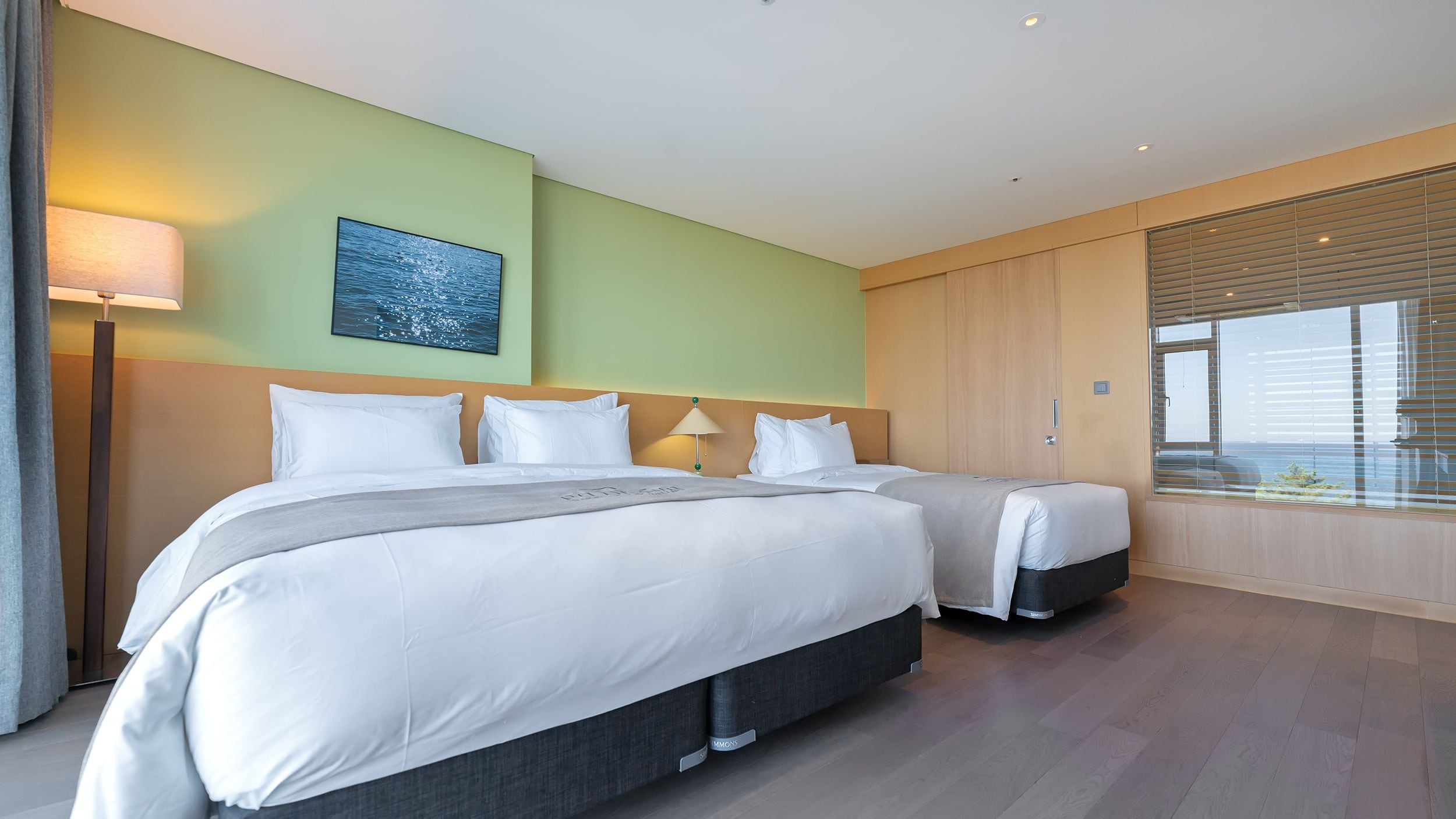 A hotel room with two beds, a large window with blinds, a painting on the wall, and a floor lamp.