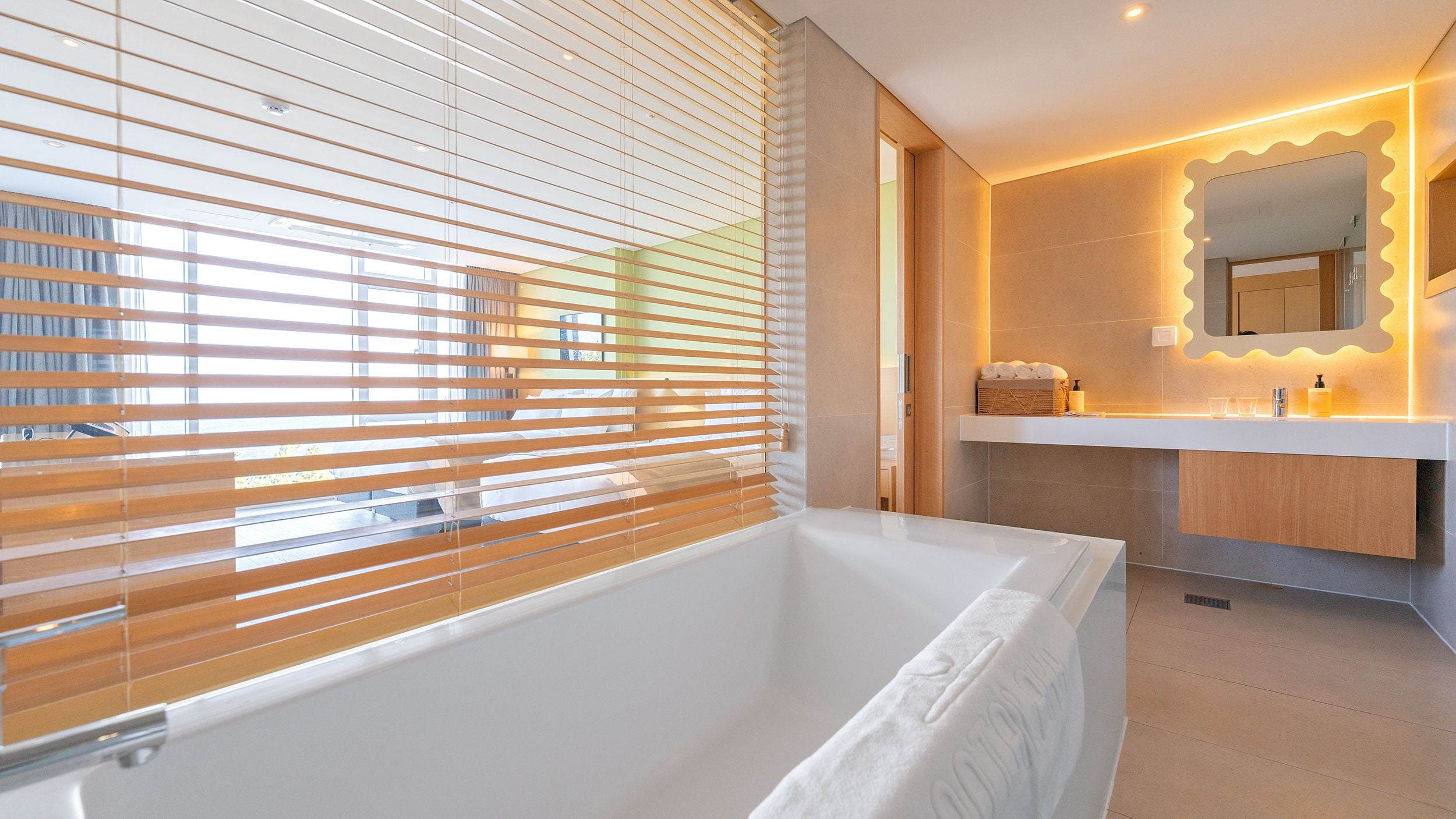 A modern bathroom with a bathtub, a vanity with a mirror, and a window with blinds.