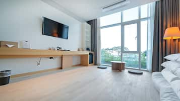 Habitación de hotel moderna con televisión de pantalla plana, escritorio con teléfono, cubo de basura, mesa redonda, sofá y ventana grande con cortinas.