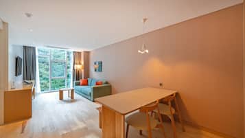 Una sala de estar moderna con un sofá, una mesa de comedor con sillas, un televisor montado en la pared y un amplio ventanal con cortinas.
