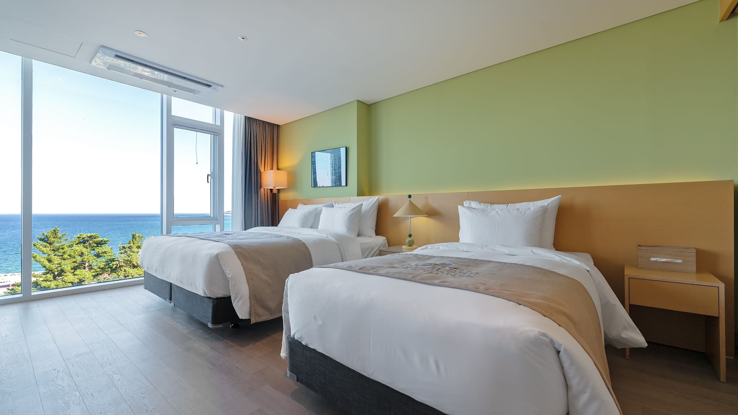 A hotel room with two beds, a large window with a view of the sea, and a nightstand with a lamp.