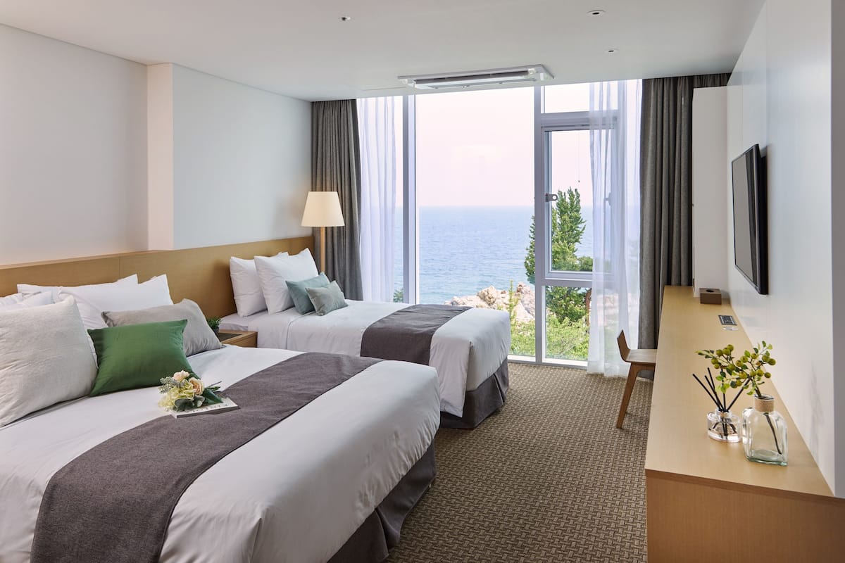 A hotel room with two beds, a desk, a TV, and a balcony with a view of the sea.