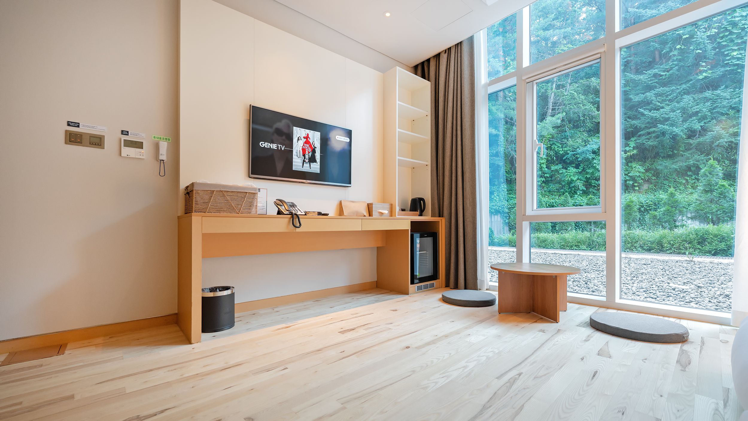 A modern room with a TV, a desk, a trash bin, and a window with a view of trees.