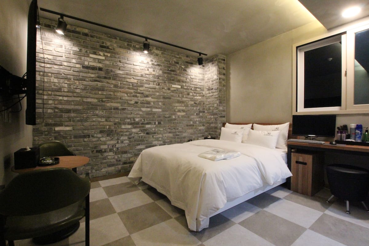 Quarto de hotel com parede de tijolos, uma cama com roupa de cama branca, uma escrivaninha com um computador e uma cadeira.