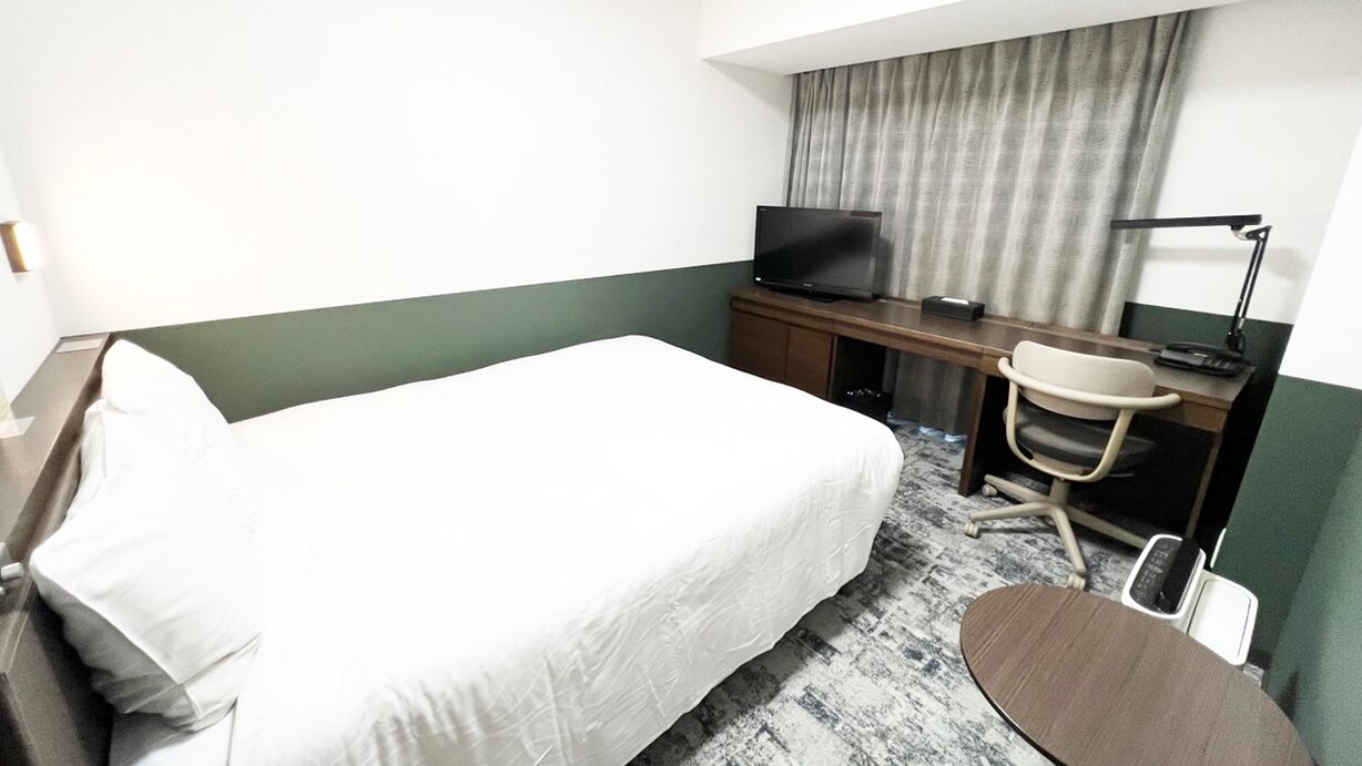Quarto de hotel com cama, escrivaninha com televisão, uma cadeira e abajur.