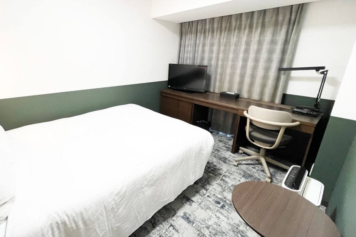 A hotel room with a bed, a desk with a television, a chair, and a desk lamp.