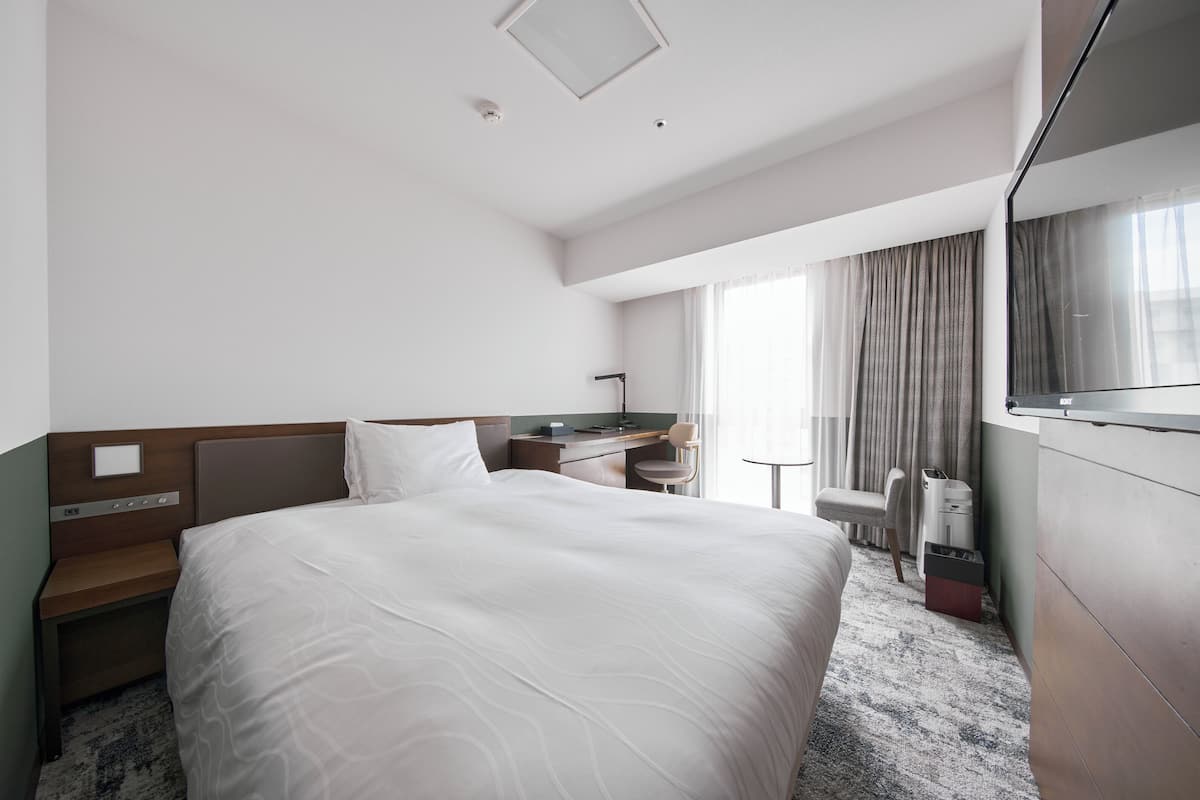 A hotel room with a large bed, a desk, a chair, a TV, and a window with curtains.