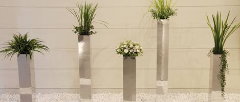 Four tall, narrow planters with plants against a plain wall, surrounded by white pebbles.
