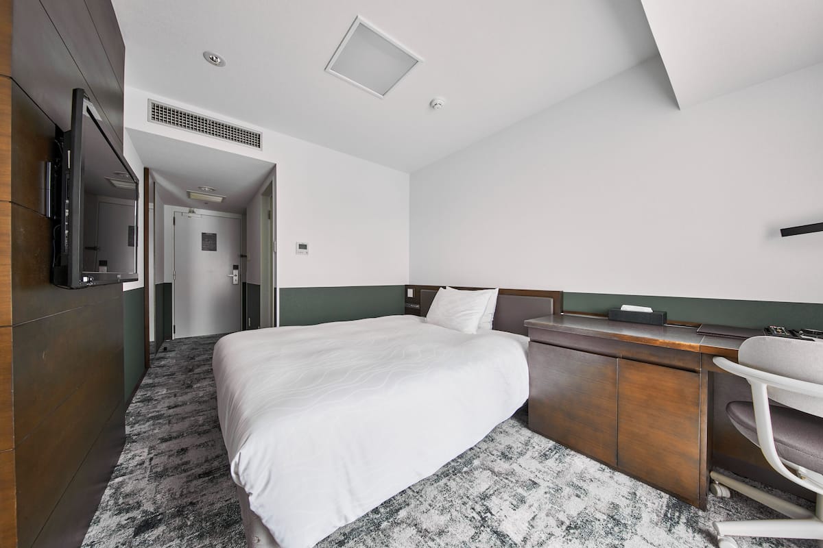 A hotel room with a large bed, a desk, a chair, a TV mounted on the wall, and a door leading to another room.