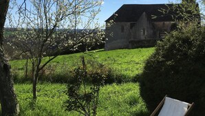 A grassy area with a wooden chair, a tree, and a building with a pointed roof in the background. - Charming 19th century country house   all comforts, great charm   true nature. (Ceaulmont)