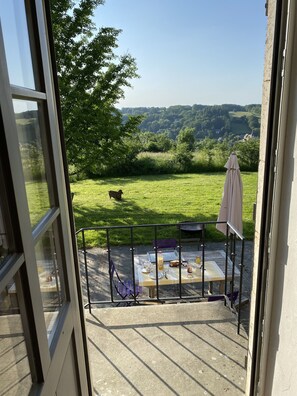 Een balkon met een eettafel, een hond op de achtergrond en uitzicht op een groen landschap.