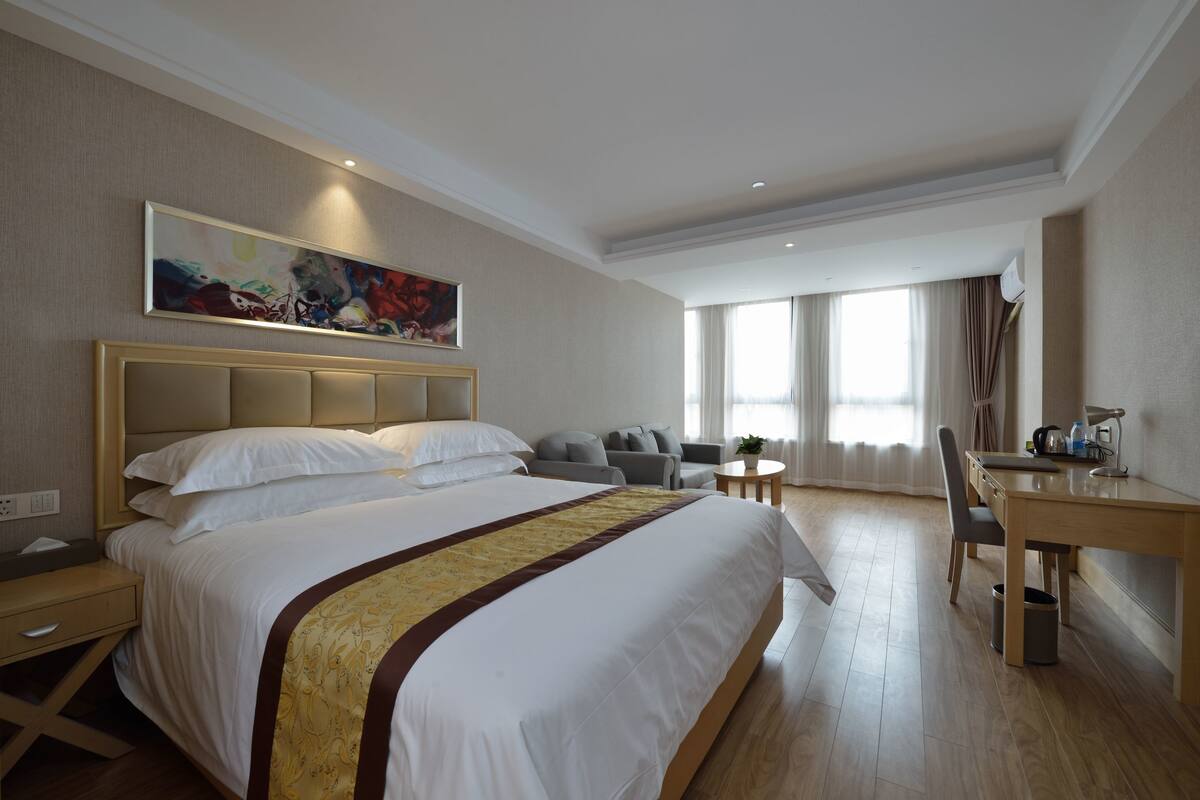 A hotel room with a large bed, a desk, a chair, a sofa, and a painting on the wall.