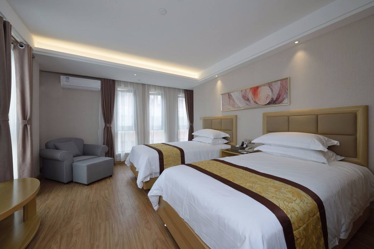 A hotel room with two beds, a sofa, a wooden floor, and an air conditioning unit.