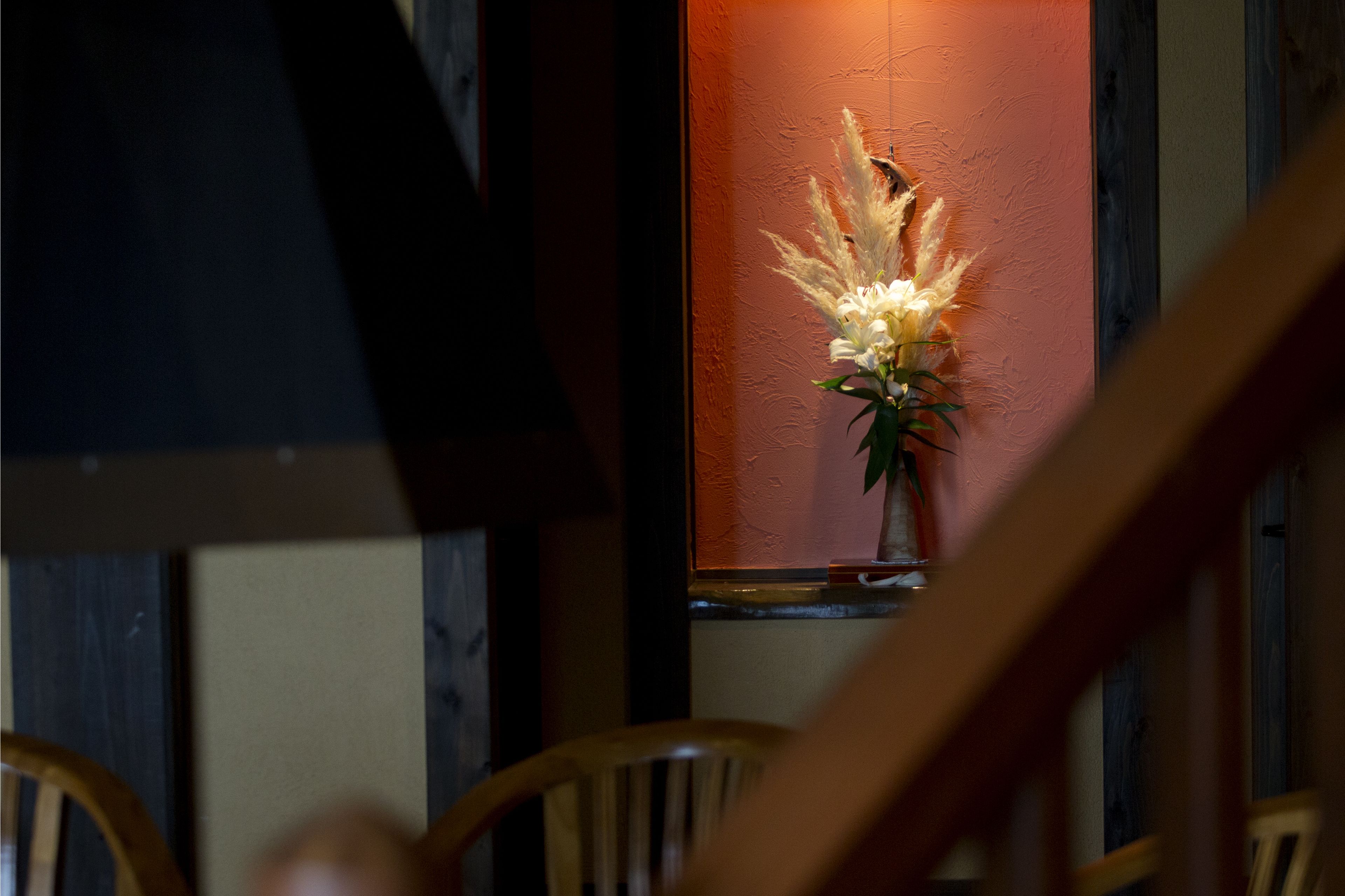 A room with a wooden chair in the foreground, a window with a vase of flowers, and a wooden wall.