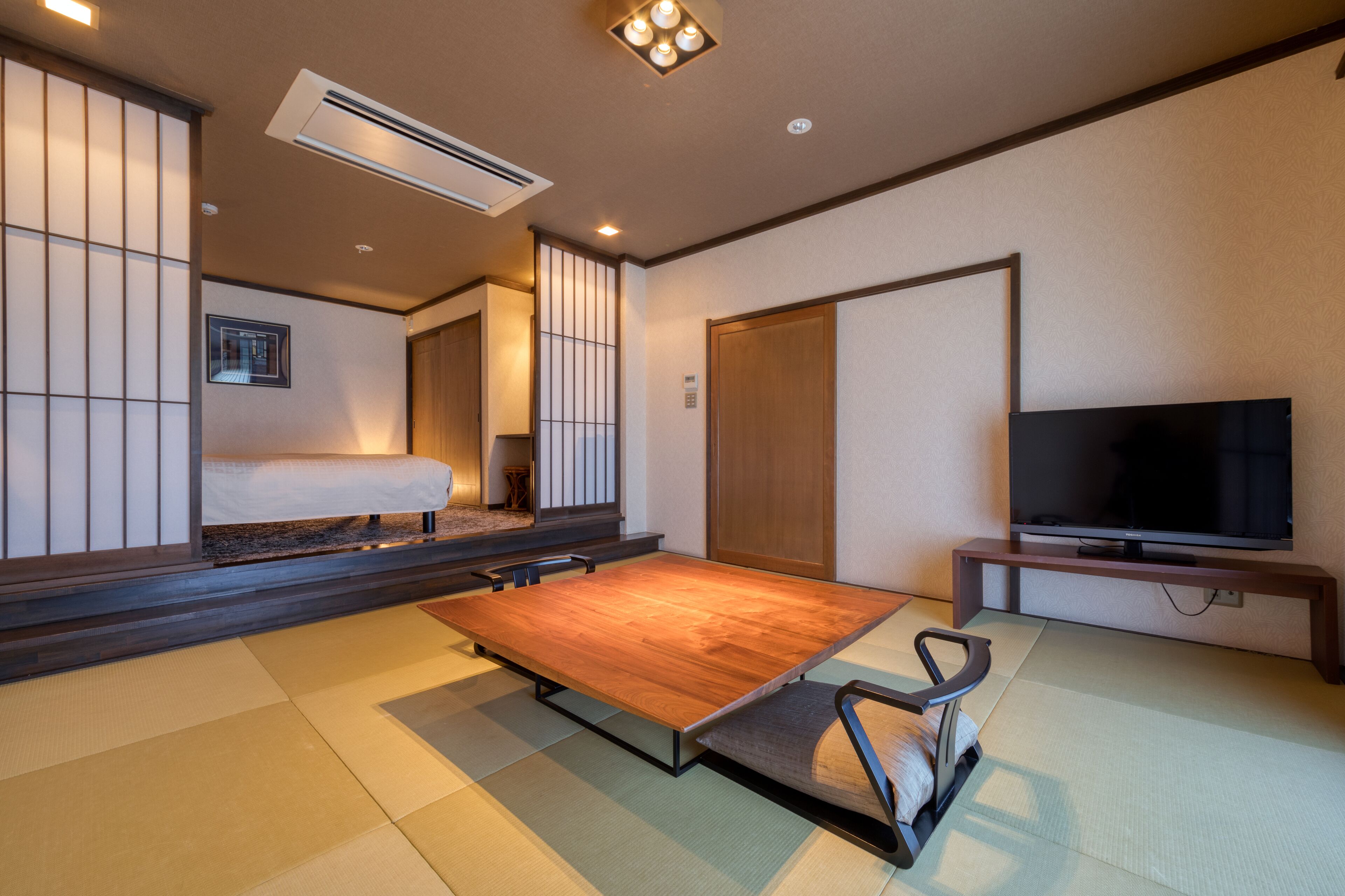 Una habitación de estilo japonés tradicional, con suelo de tatami, una cama baja de madera, un televisor sobre un mueble y puertas correderas.