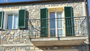 A two-story stone building with green shutters, a balcony with metal railings, and a large sliding glass door.