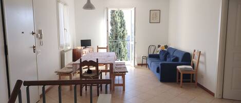 A spacious living area with a dining table, chairs, a sofa, and a balcony with a view of greenery.