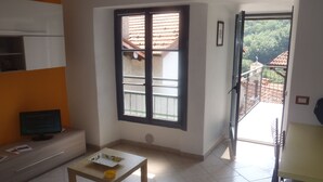 Una habitación con ventana, una mesa con bandeja y un tazón amarillo, un televisor y una puerta que da a un balcón al aire libre.