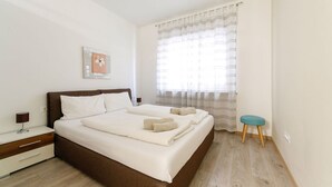 Un dormitorio con una cama grande, una mesita de noche, una pequeña silla y un ventanal con cortinas translúcidas.