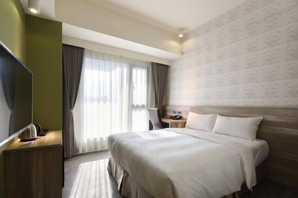 Hotellihuone, jossa on suuri sänky, televisio, työpöytä lampun kanssa ja ikkunasta avautuva näkymä ulos.