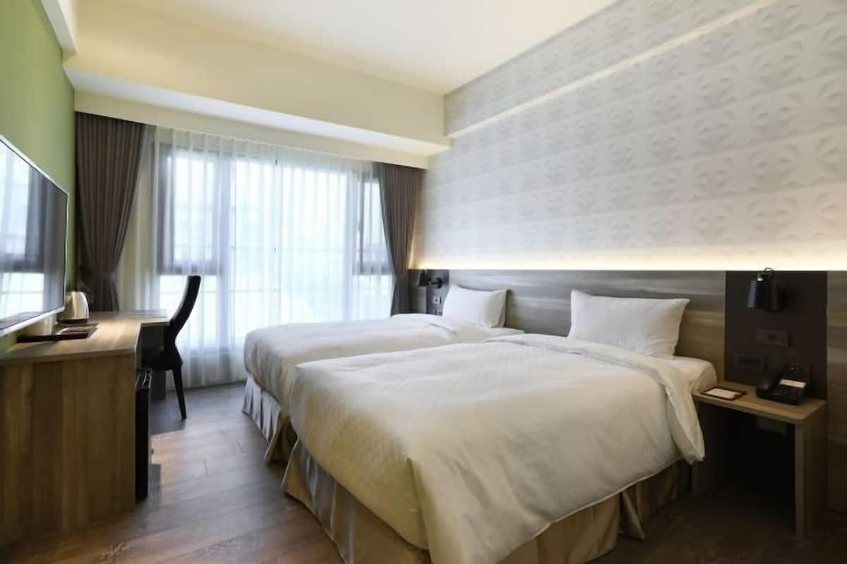 Hotellihuone, jossa on kaksi sänkyä, työpöytä, tuoli, televisio ja suuri ikkuna, jossa on verhot.