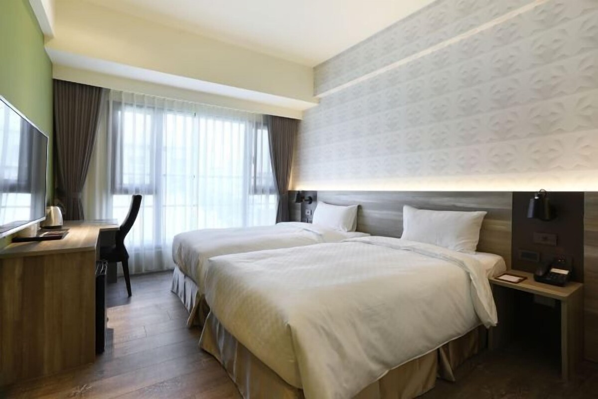 A hotel room with two beds, a desk, a chair, a TV, and a large window with curtains.