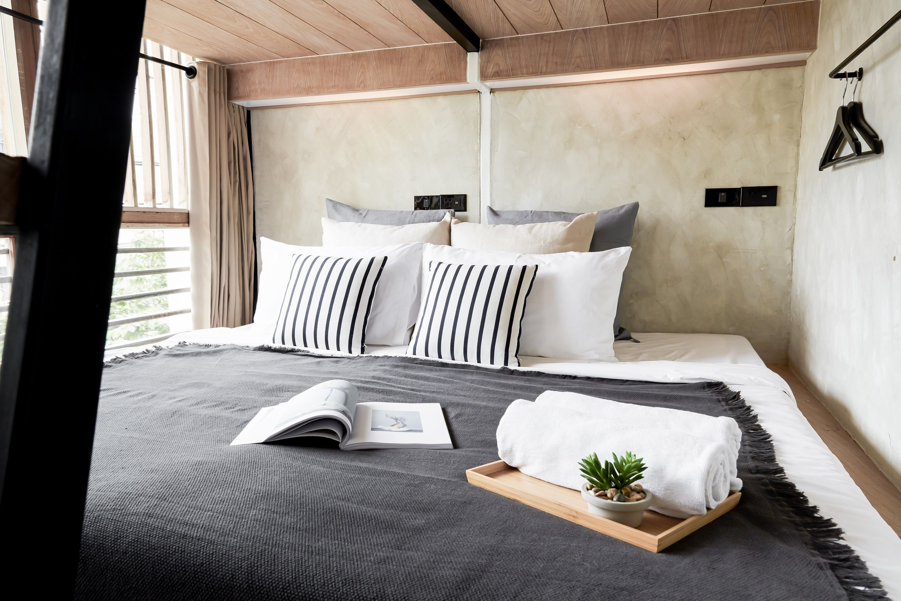 A modern bedroom with a large bed, striped pillows, a bedside table with a book and a plant, and a view of the outdoors.