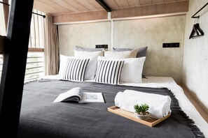 A modern bedroom with a large bed, striped pillows, a bedside table with a book and a plant, and a view of the outdoors. - Vann Bangkok Boutique House (Bangkok)