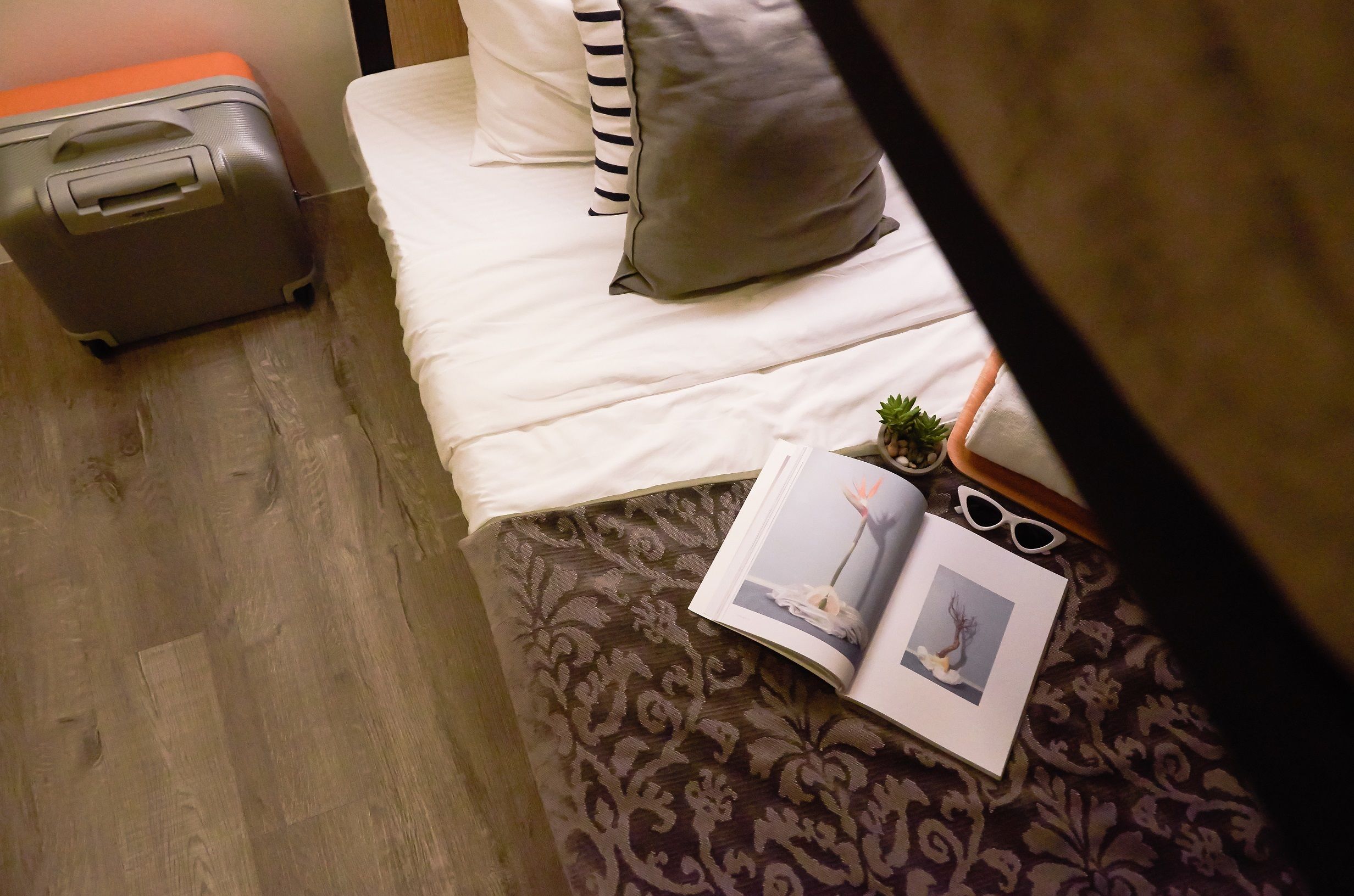 Un letto con copriletto decorato, un cuscino, una valigia, un libro e un paio di occhiali da sole sopra il letto.