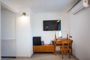 A compact room with a wooden desk, a television mounted on the wall, a small refrigerator, and a radiator.