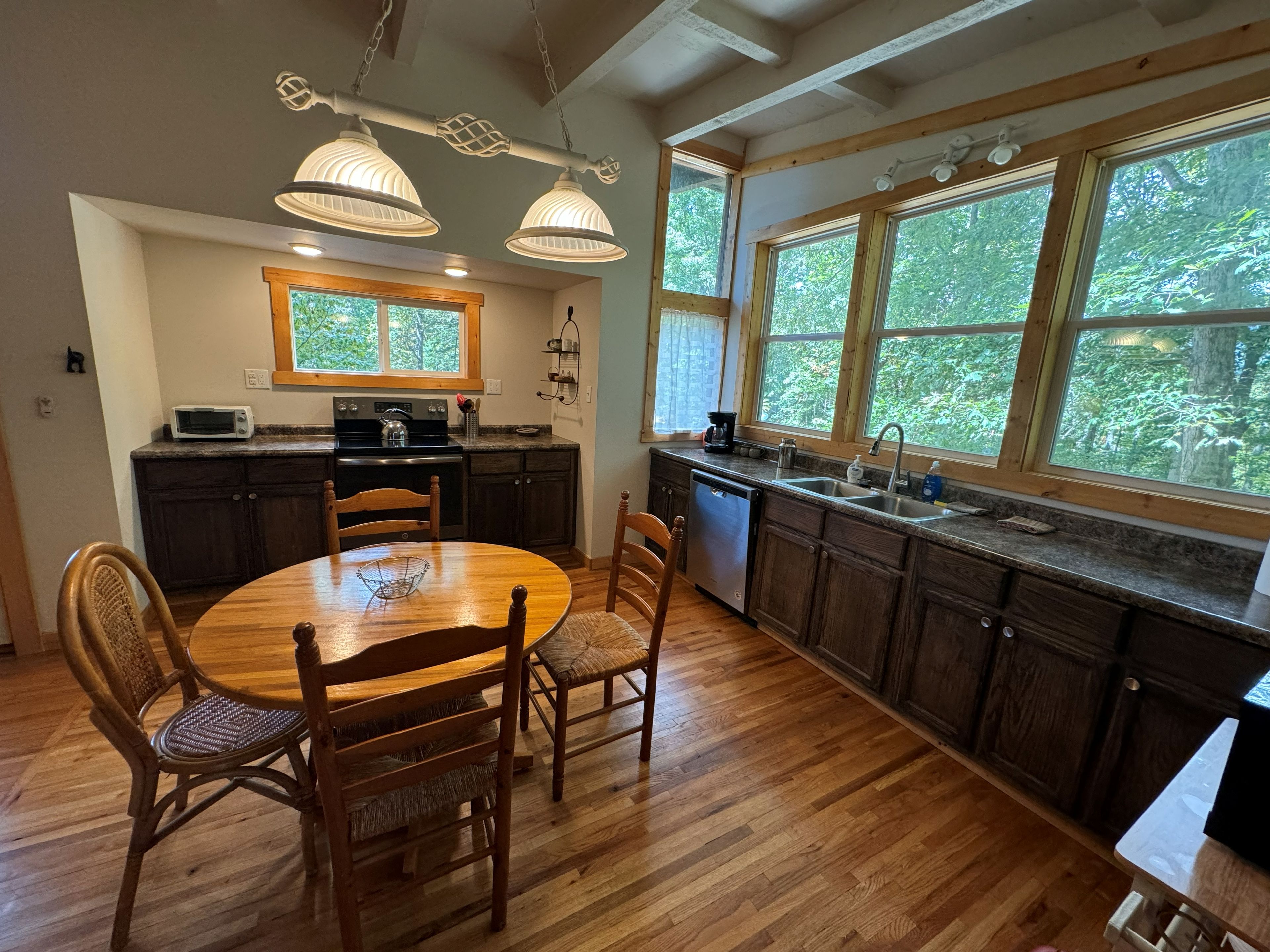 A kitchen with wooden cabinets, a dining table with chairs, and large windows offering a view of greenery.
