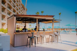 An outdoor bar area with a wooden counter, bar stools, and a view of the beach and palm trees. - Sonoran Spa W107 - Ground-Floor Beachfront Condo,  Steps to Pool & Beach (Puerto Peñasco)