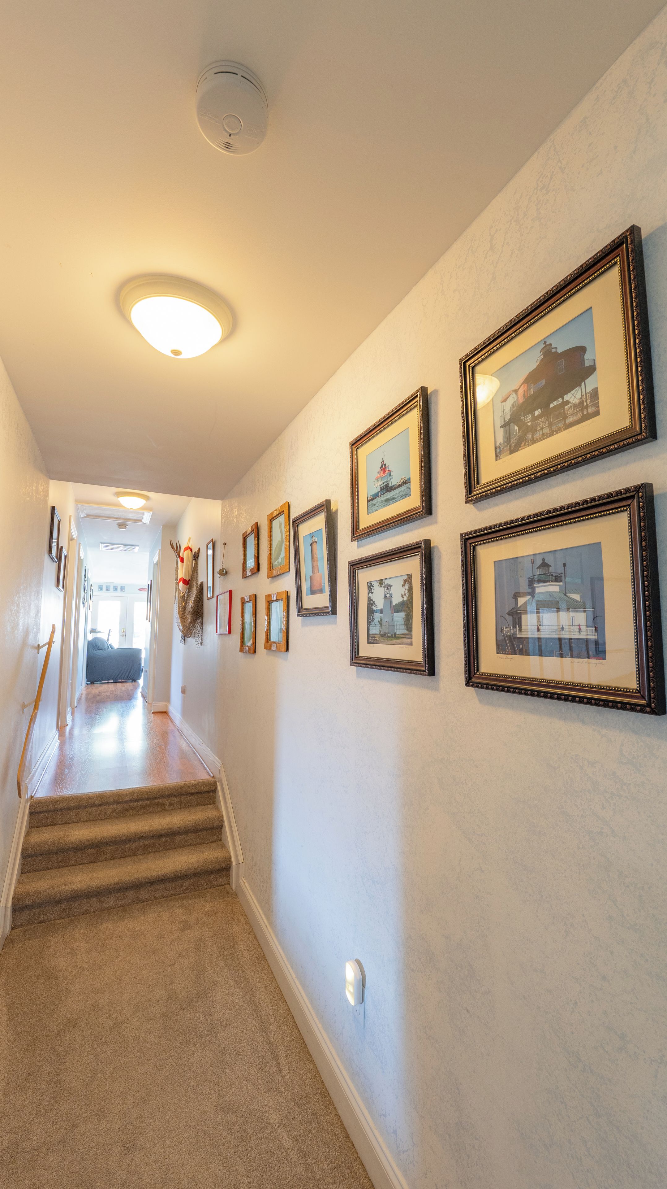 Un couloir avec des tableaux encadrés accrochés au mur, un escalier recouvert de moquette et un luminaire au plafond.
