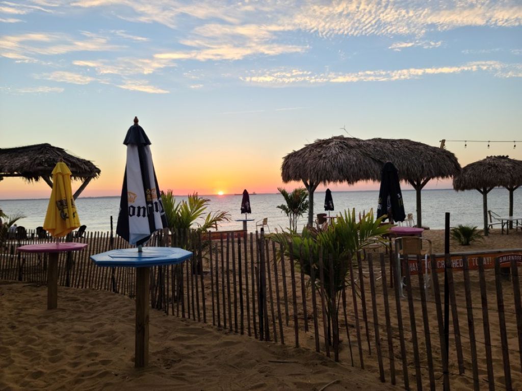 A beach with thatched umbrellas and tables, a wooden fence, and a sunset in the background.